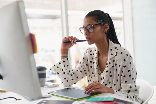 A focused professional wearing stylish glasses and a polka dot blouse contemplates a creative project on her computer in a modern office setting.