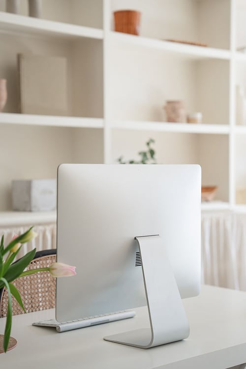 A minimalist home office setup featuring a sleek computer on a clean white desk, enhanced by a vibrant tulip and a backdrop of stylish shelves.