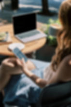 A person relaxes at an outdoor café, sipping on coffee and browsing their phone, with a laptop and a plate of food on the table, enjoying the sunny day.
